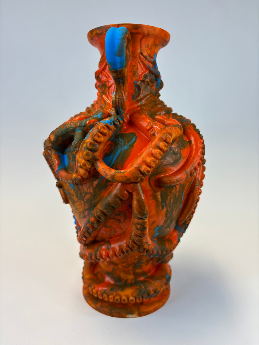 An intricately carved vase with a textured, organic pattern in terracotta orange, dark brown, and bright blue accents, shown against a plain light gray background.