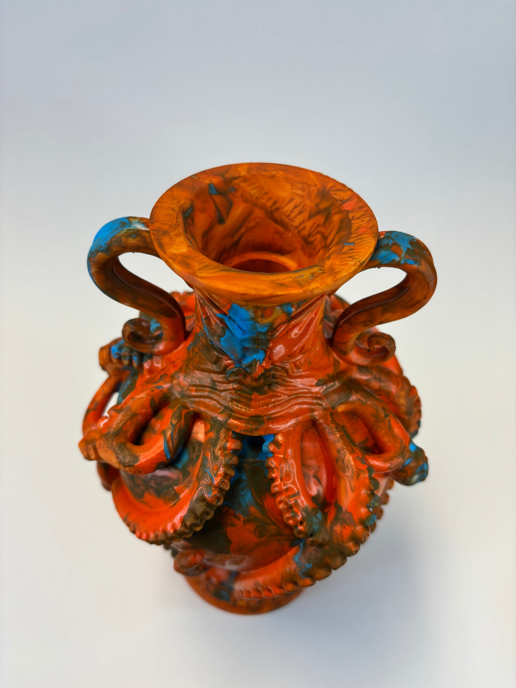 An ornate sculptural vase with a flared rim and two handles, featuring a highly detailed octopus design in a distressed orange-red glaze with dark brown and bright blue accents, set against a plain light gray background.