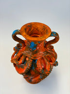 An ornate sculptural vase with a flared rim and two handles, featuring a highly detailed octopus design in a distressed orange-red glaze with dark brown and bright blue accents, set against a plain light gray background.