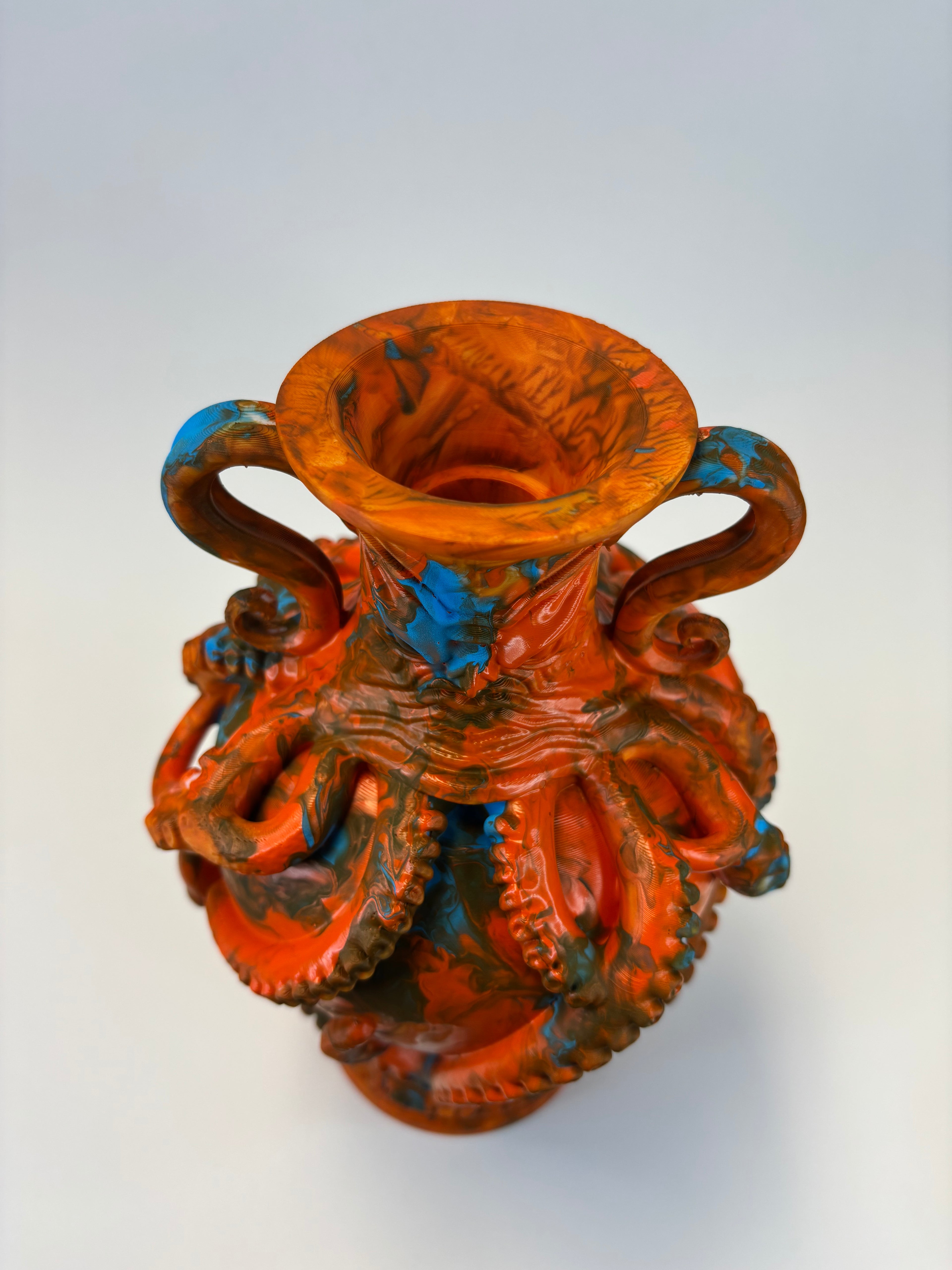 An ornate sculptural vase with a flared rim and two handles, featuring a highly detailed octopus design in a distressed orange-red glaze with dark brown and bright blue accents, set against a plain light gray background.