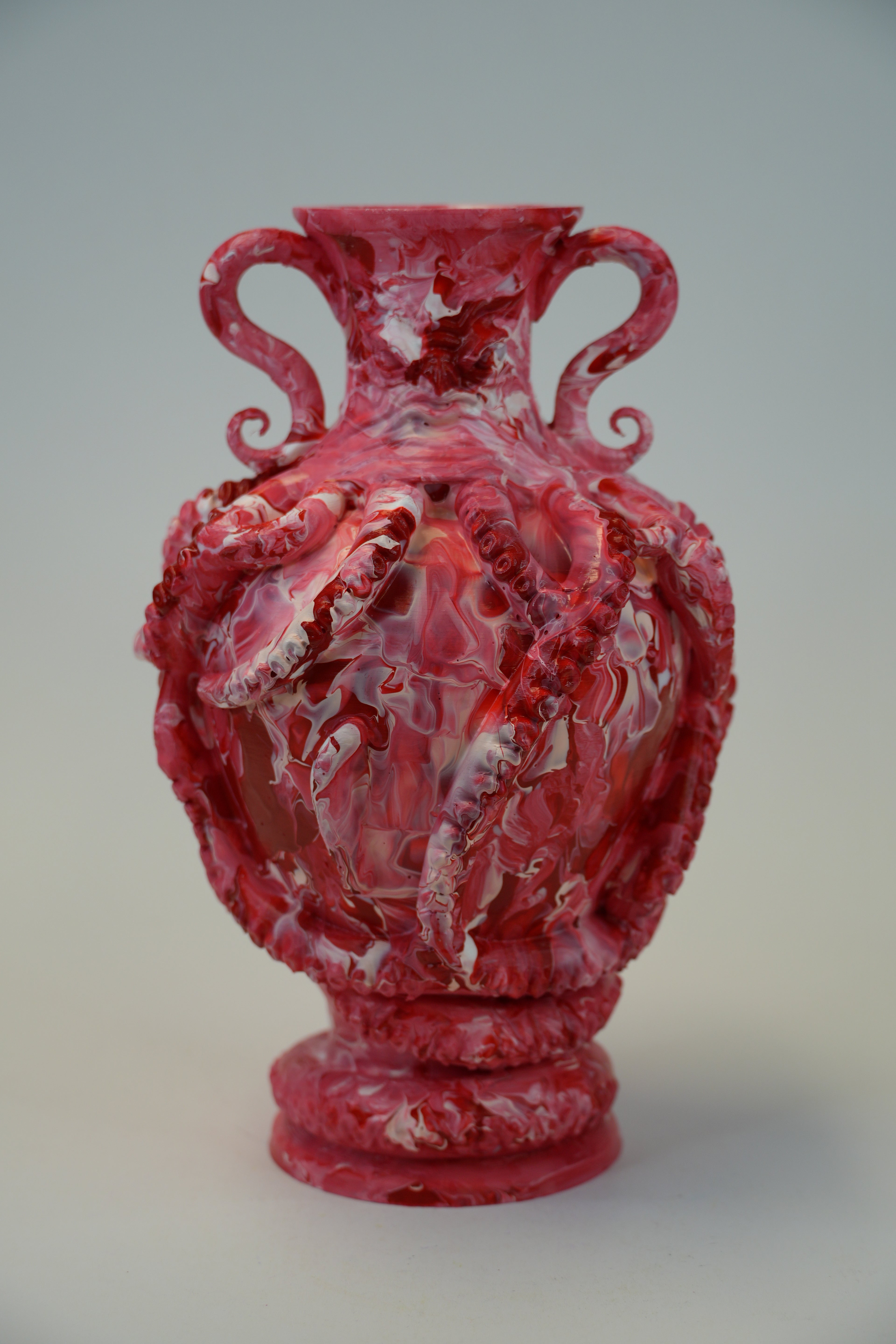 A red and white marbled resin sculpture of a classical urn with twisted handles, photographed against a neutral light gray background.
