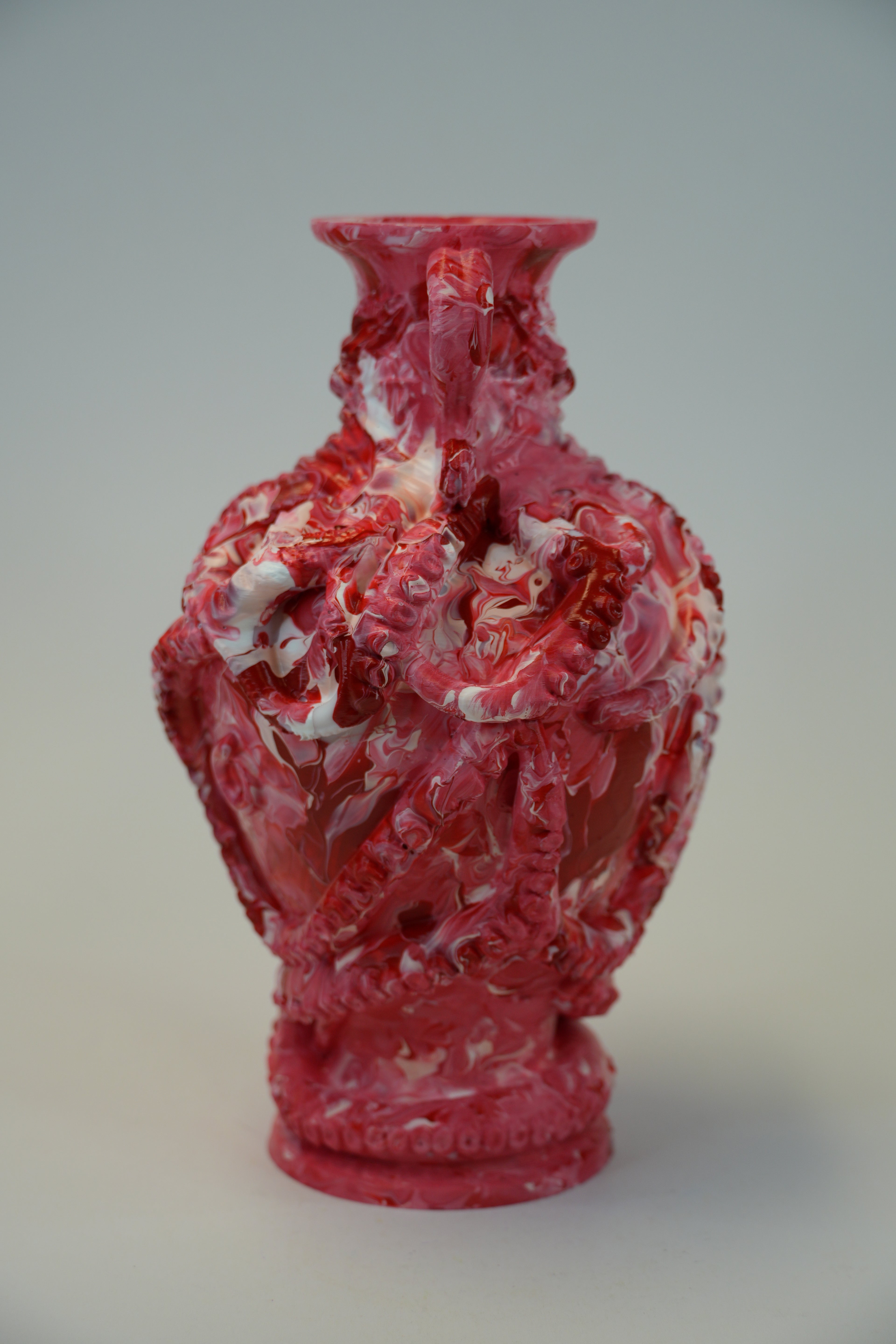 A decorative ceramic vase with an intricate, raised floral and scroll pattern in a vibrant pinkish-red glaze with white marbled accents, set against a neutral light gray background.