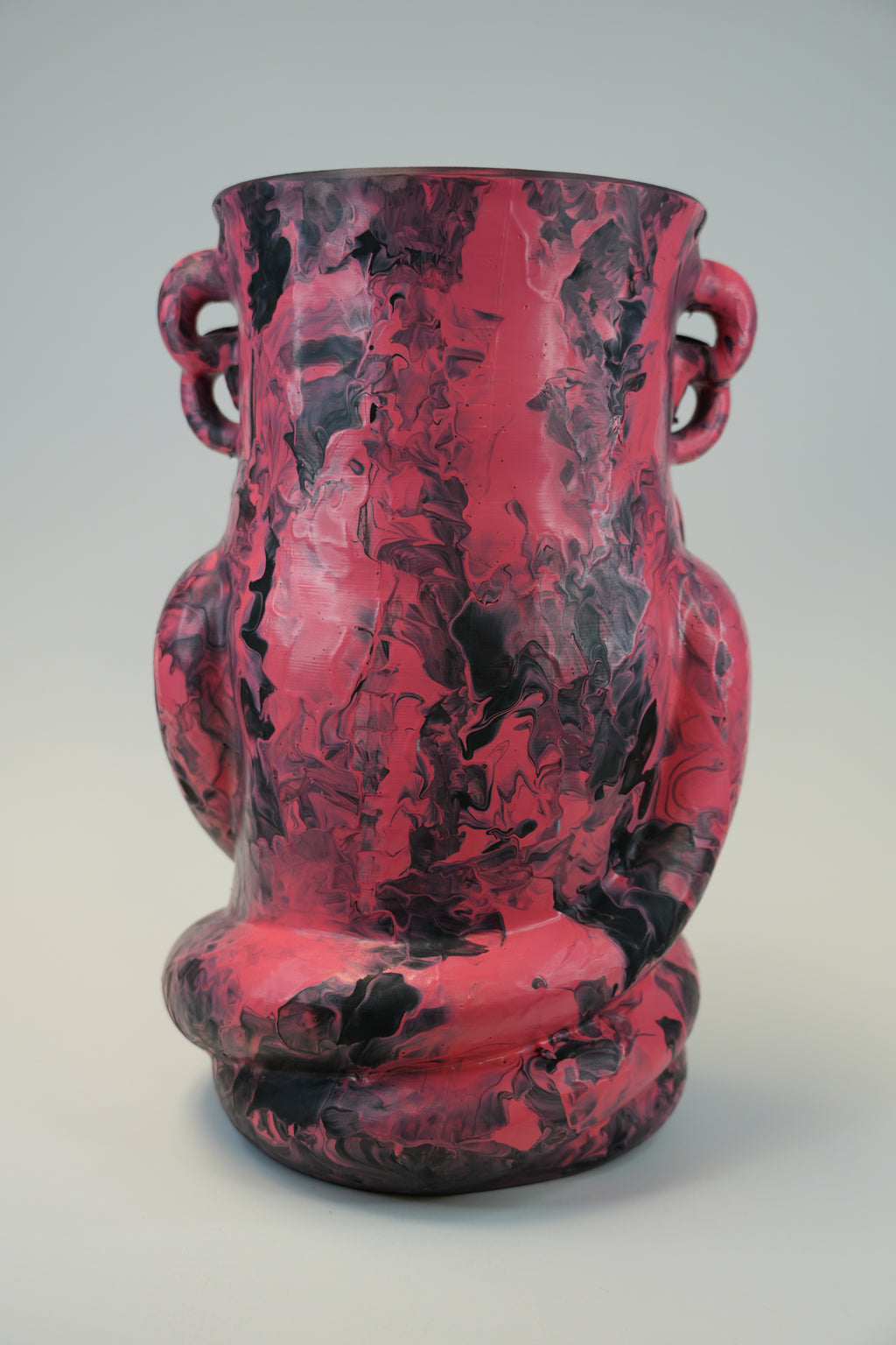 A sculptural ceramic vase with a glossy pink and black marbled pattern, featuring integrated side handles and a coiled lower section, photographed against a neutral light gray background.
