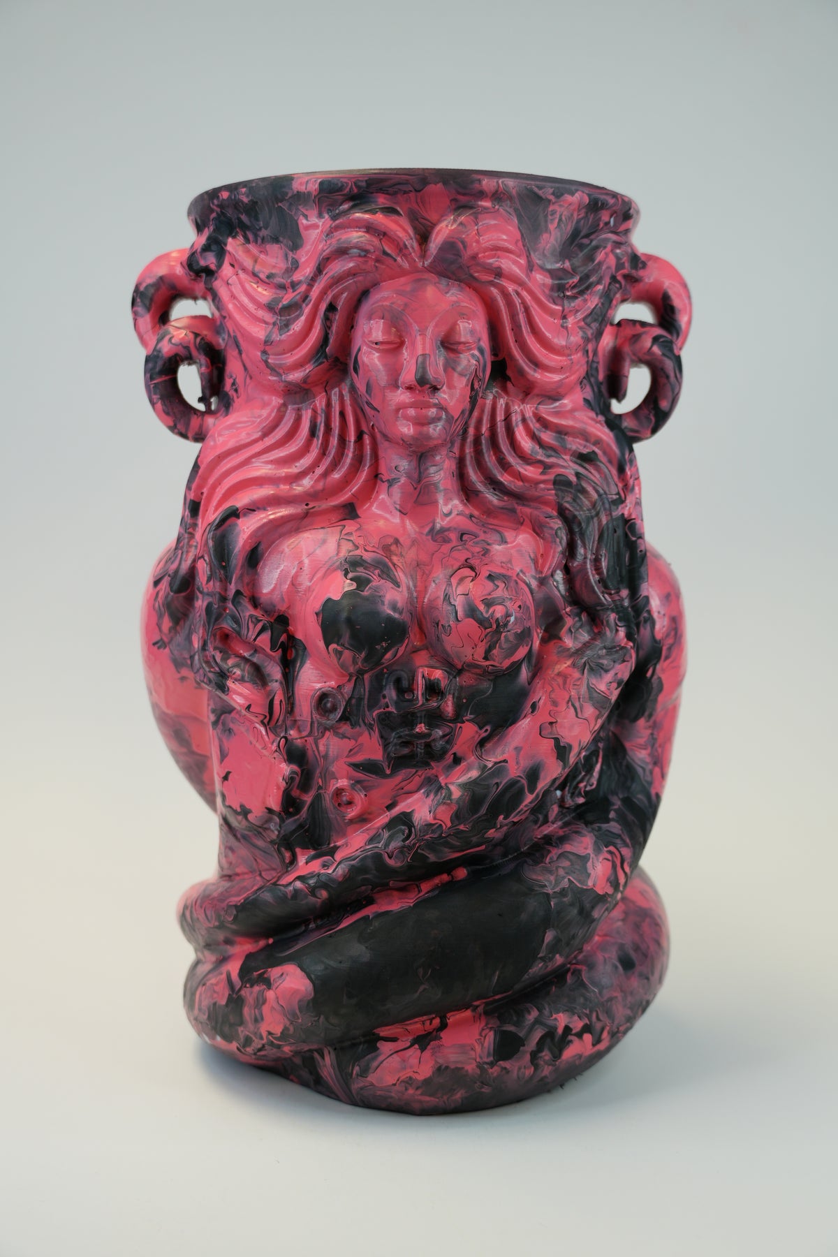 A sculptural ceramic vase featuring a profile figure with a coiled body, rendered in a marbled mix of deep pink, magenta, and black glaze on a light gray studio background.