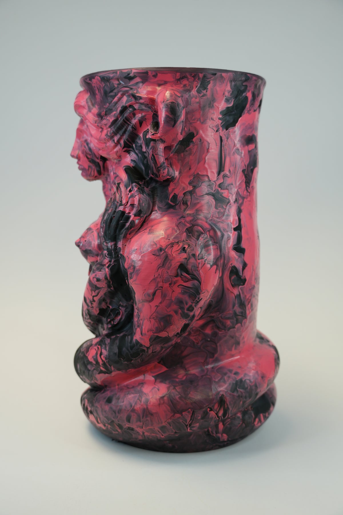 A sculptural ceramic vase featuring a profile figure with a coiled body, rendered in a marbled mix of deep pink, magenta, and black glaze on a light gray studio background.