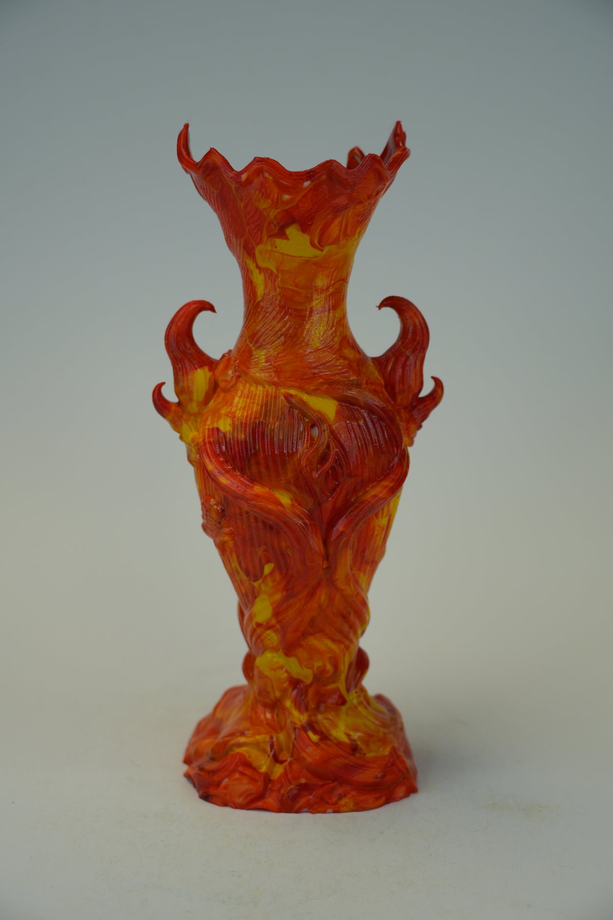 A sculptural glass vase with a flared rim and two side handles, crafted in a marbled orange, red, and yellow glass style, set against a neutral gray background.