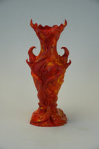 An ornate sculptural vase with a flared rim, featuring a textured, swirling pattern in vibrant red and orange with yellow highlights, set against a neutral light gray background.