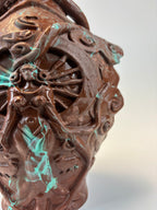 Close-up of an intricately carved brown sculptural object with swirling organic patterns and small turquoise-green marbled accents, shown against a plain light background.
