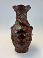A dark brown carved decorative object with a textured, organic surface and subtle turquoise patina highlights, shown against a plain light gray background.