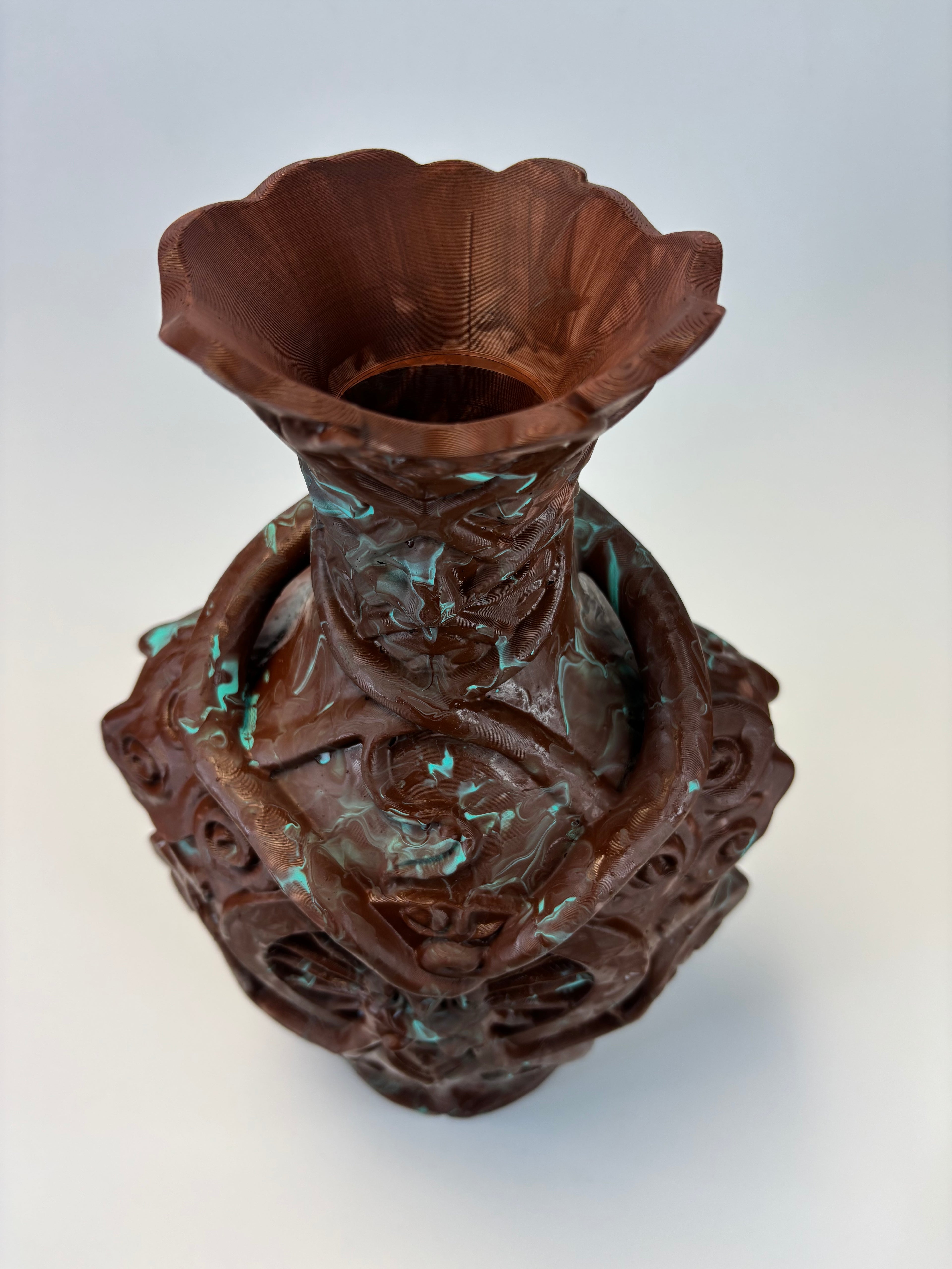 An ornate sculptural vase with a flared trumpet-like rim, featuring intricate relief carvings and a patinated dark brown finish with subtle turquoise-green accents, shown on a light gray background.