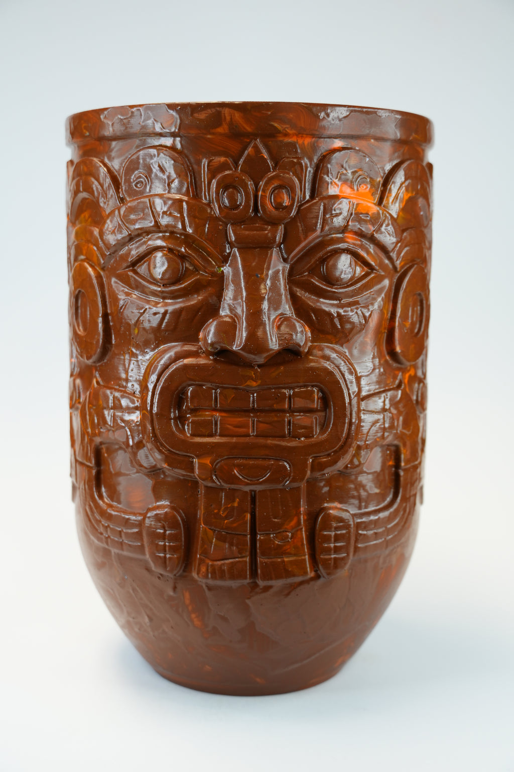 A carved brown ceramic or stone vessel with a detailed face motif, photographed against a plain light gray background.