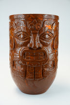 A carved brown ceramic or stone vessel with a detailed face motif, photographed against a plain light gray background.