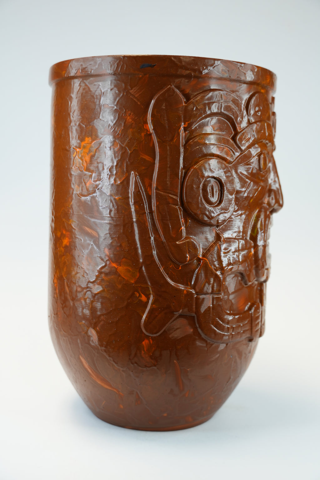 A brown translucent ceramic cup with a carved Aztec skull design on the side, photographed against a light gray background.