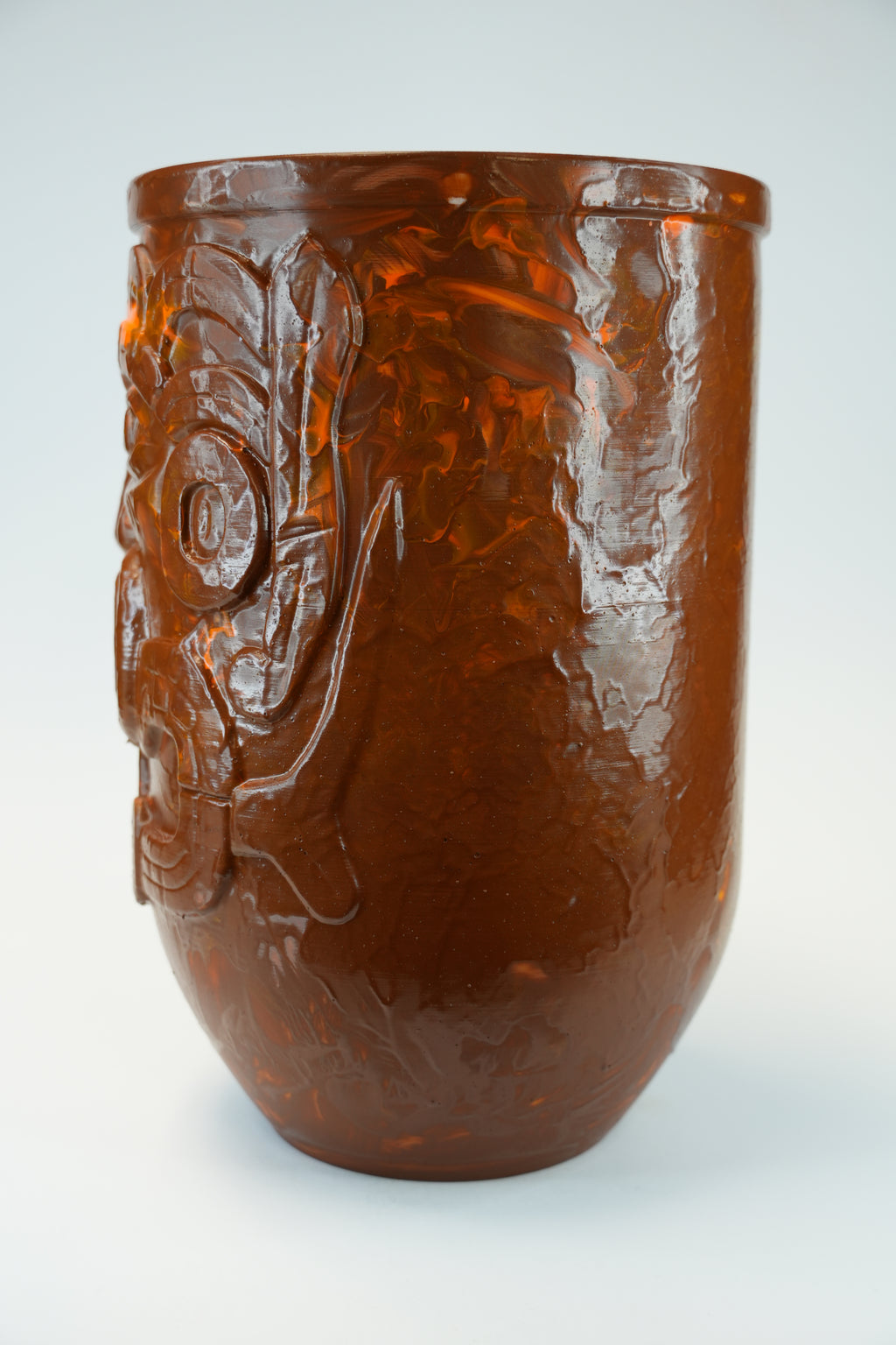 An amber-brown translucent glass cup with a raised relief design of a stylized face on the side, shown against a light gray background.