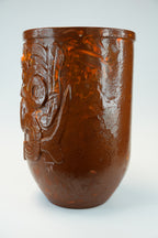 An amber-brown translucent glass cup with a raised relief design of a stylized face on the side, shown against a light gray background.