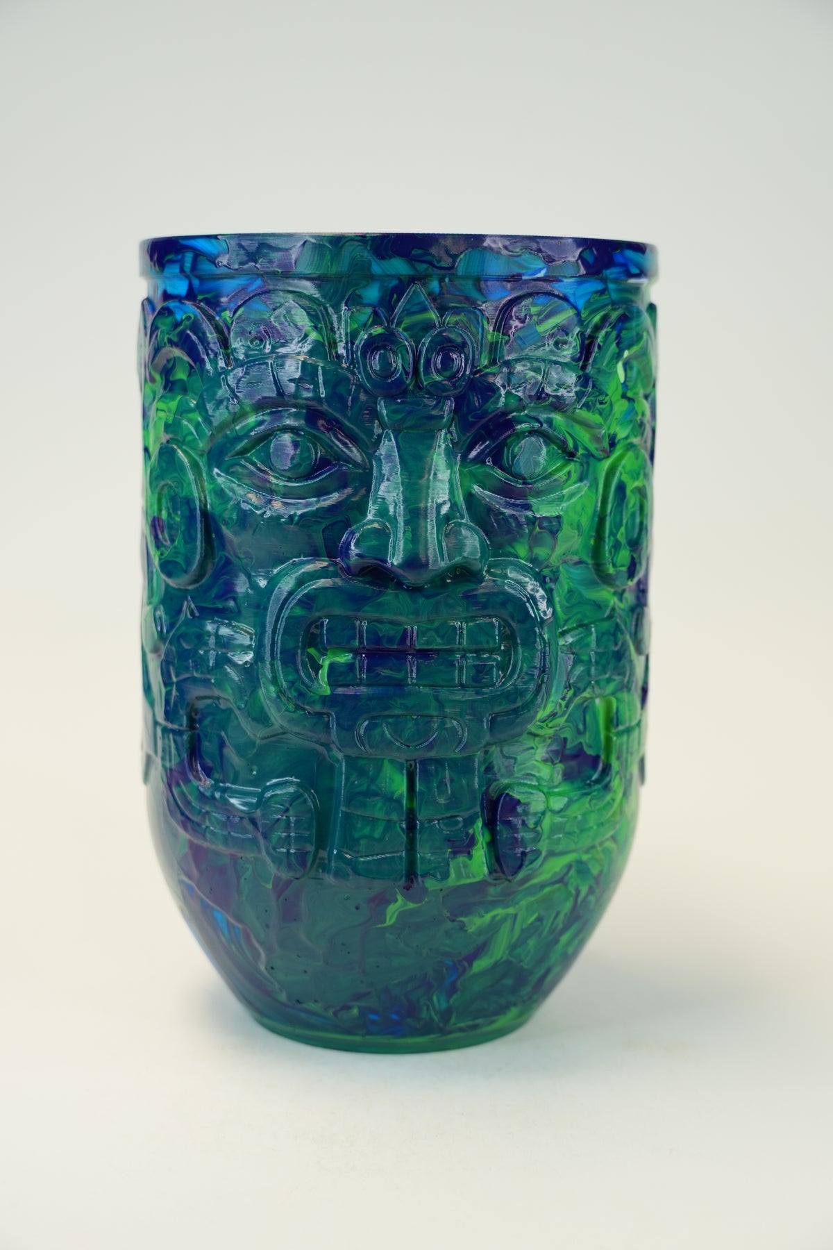 A sculptural tumbler vase featuring an intricate Aztec-style face design, crafted from marbled blue and green glass on a light gray background.