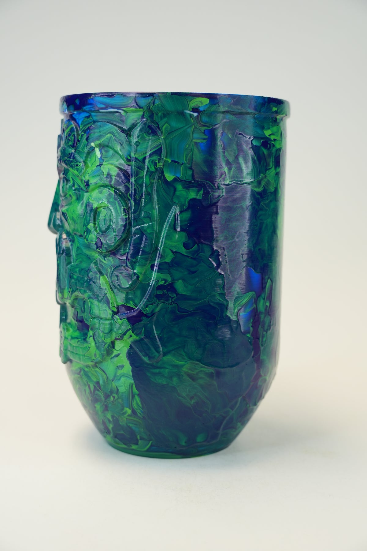 A sculptural, glossy ceramic cup or small vase with a raised face motif, featuring marbled green, blue, and dark purple glaze on a light gray background.