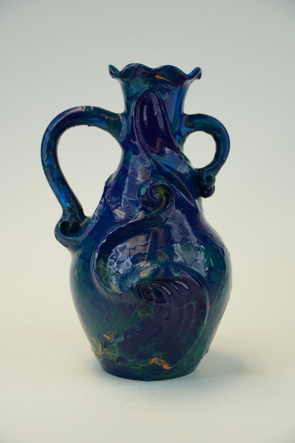 A cobalt blue glazed ceramic pitcher with two handles and a decorative embossed swirl pattern, photographed against a neutral light gray background.