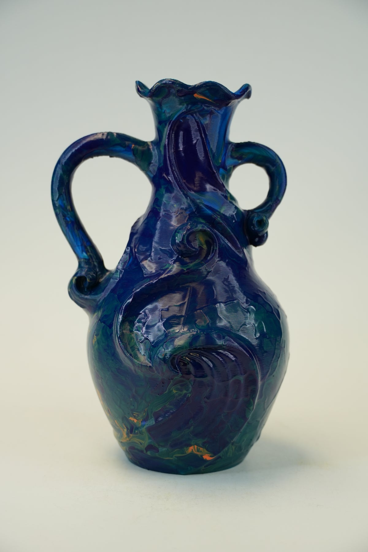 A cobalt blue glazed ceramic pitcher with two handles and a decorative embossed swirl pattern, photographed against a neutral light gray background.