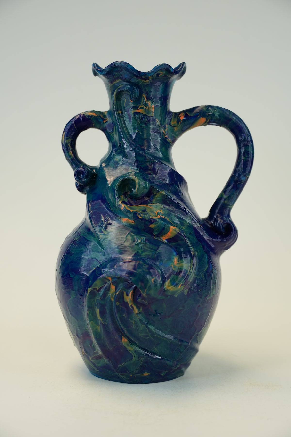 A decorative ceramic pitcher with a sculpted, organic form and two loop handles, featuring a glossy marbled glaze in deep blue, purple, and green with subtle gold-orange veining, set against a plain light gray background.