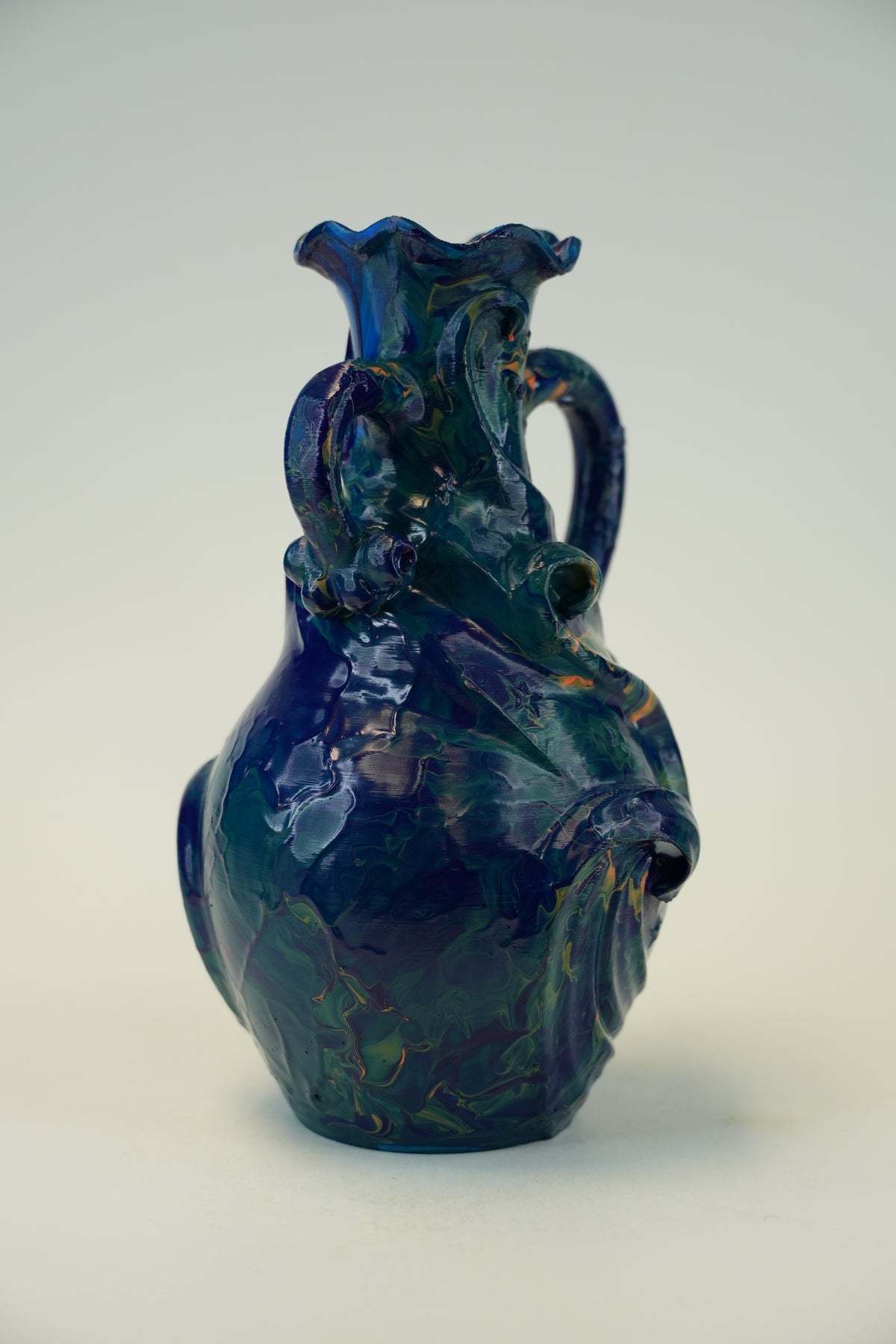 A decorative ceramic pitcher with a glossy, marbled deep blue and green glaze, featuring a wide flared rim, a side handle, and a small pouring spout, set against a plain light gray background.