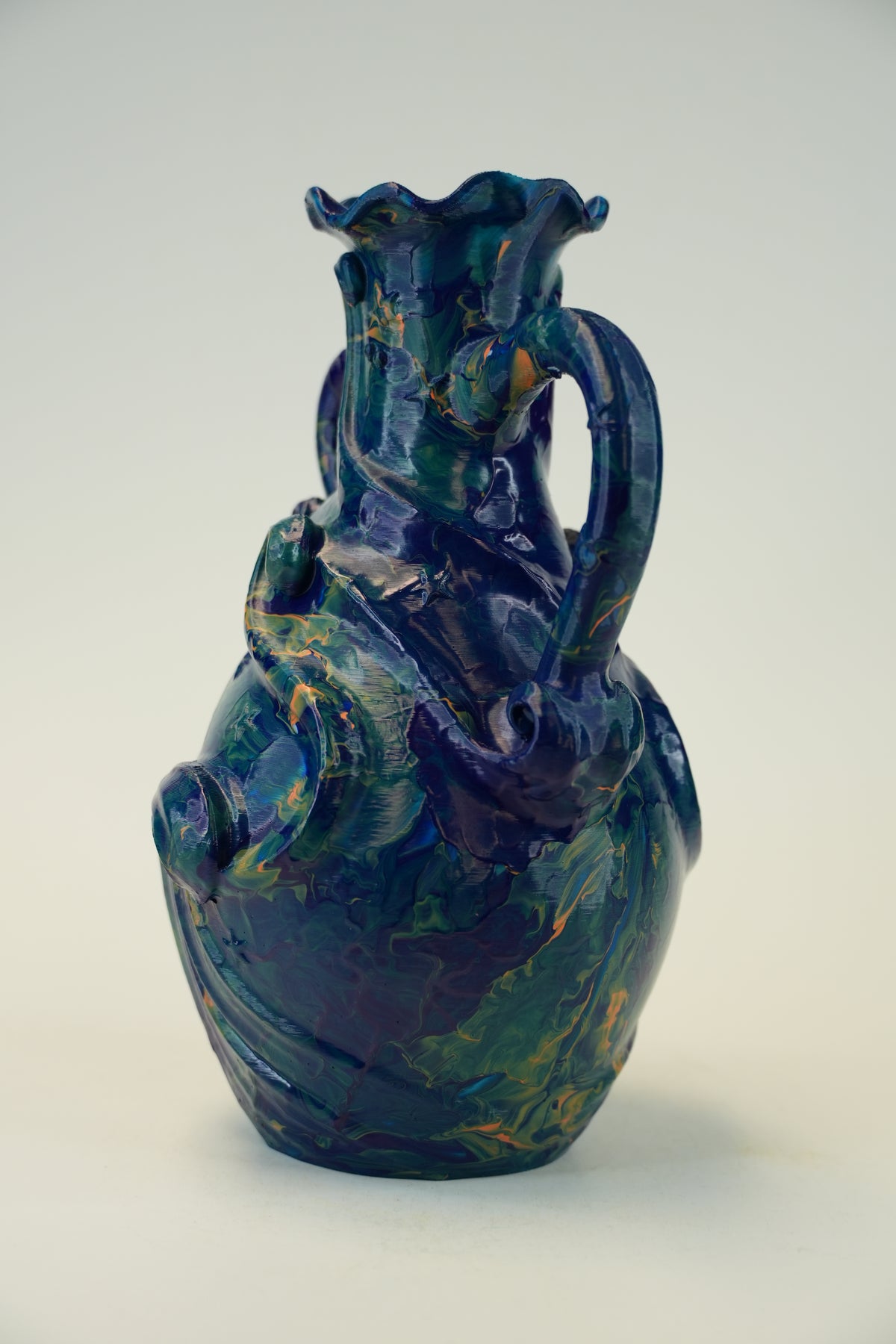 A decorative ceramic vase with a glossy, marbled glaze in deep blue, green, and gold tones, featuring a flared rim, a curved handle, and a sculpted body, set against a plain light background.
