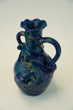 A glossy ceramic pitcher with a deep blue and green marbled glaze, featuring two handles and a flared rim, set against a plain light background.