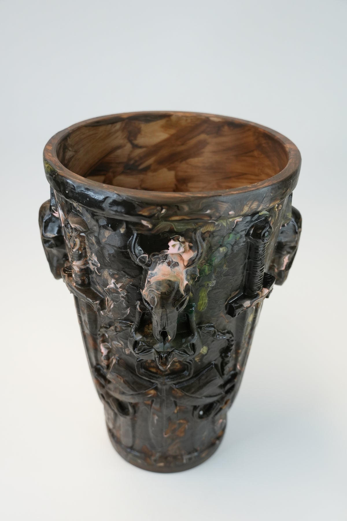 An ornate dark brown ceramic vase with a glossy finish, featuring sculpted relief details of winged cherubs and animal heads on the sides, set against a plain light gray background.