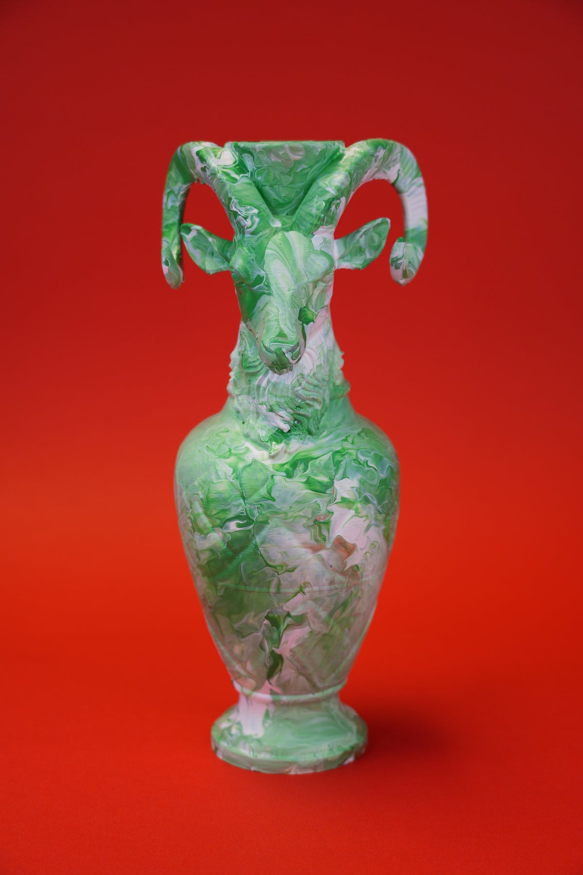 A green and white marbled ceramic ram-shaped vase with curved horns, set against a solid red background.