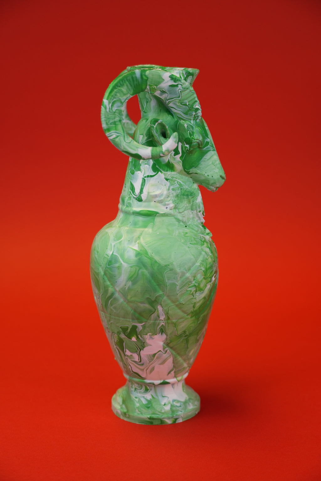 A green glass vase with a sculpted ram head handle and foot, featuring marbled green and white patterns, set against a solid red background.