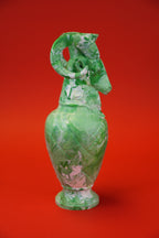 A green glass vase with a sculpted ram head handle and foot, featuring marbled green and white patterns, set against a solid red background.