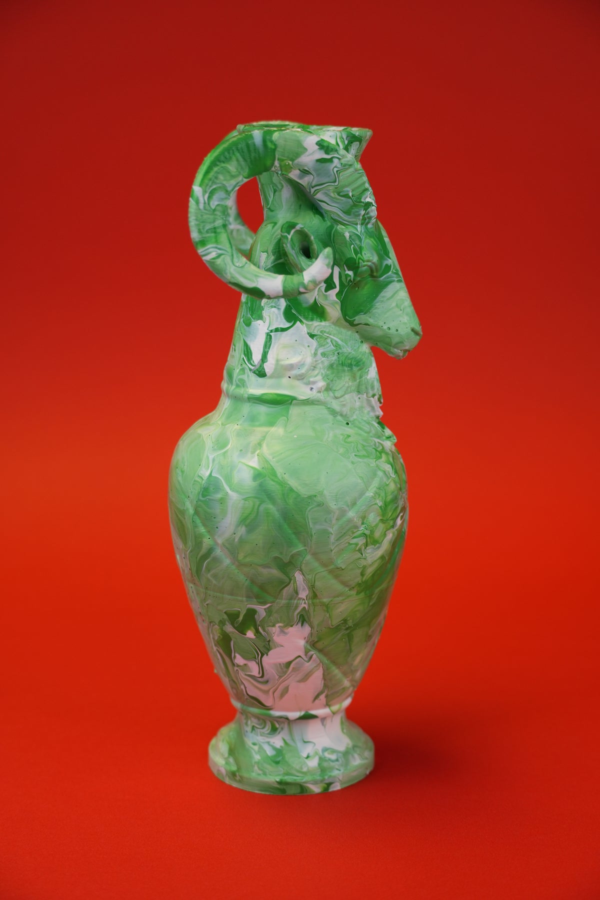 A green glass vase with a sculpted ram head handle and foot, featuring marbled green and white patterns, set against a solid red background.