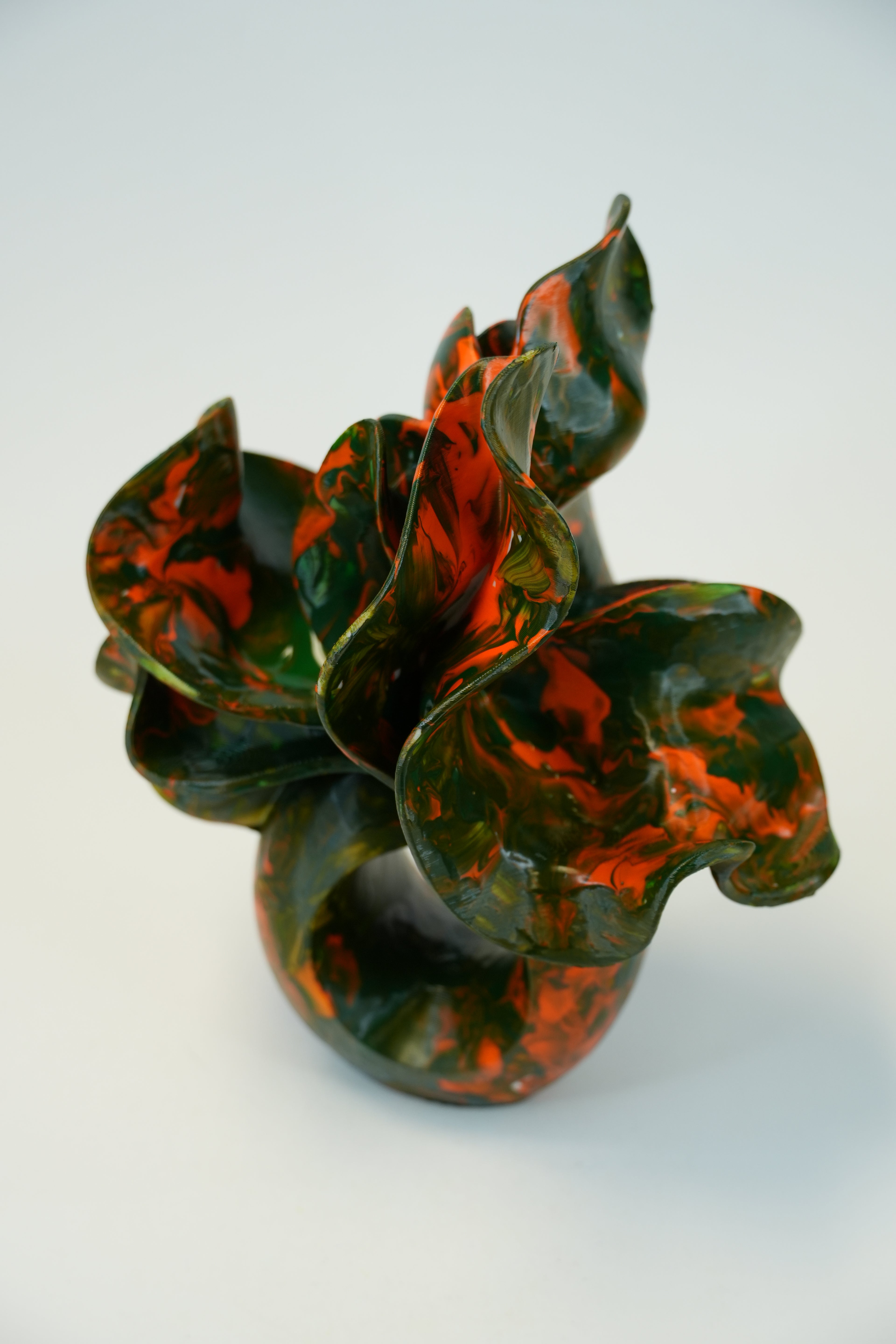 A sculptural ceramic vase with organic, leaf-like ruffled petals in glossy dark green and burnt orange marbled glaze, set against a plain light gray background.