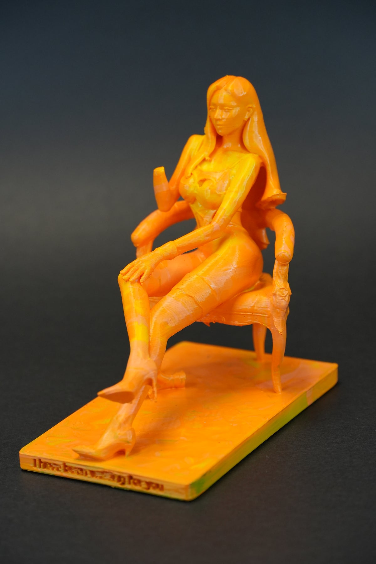 An orange figurine of a seated woman on a matching rectangular base, photographed against a dark gray gradient background.