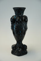 A black gothic-style ceramic vase decorated with sculpted skulls and dark blue marbled accents, set against a neutral light gray background.