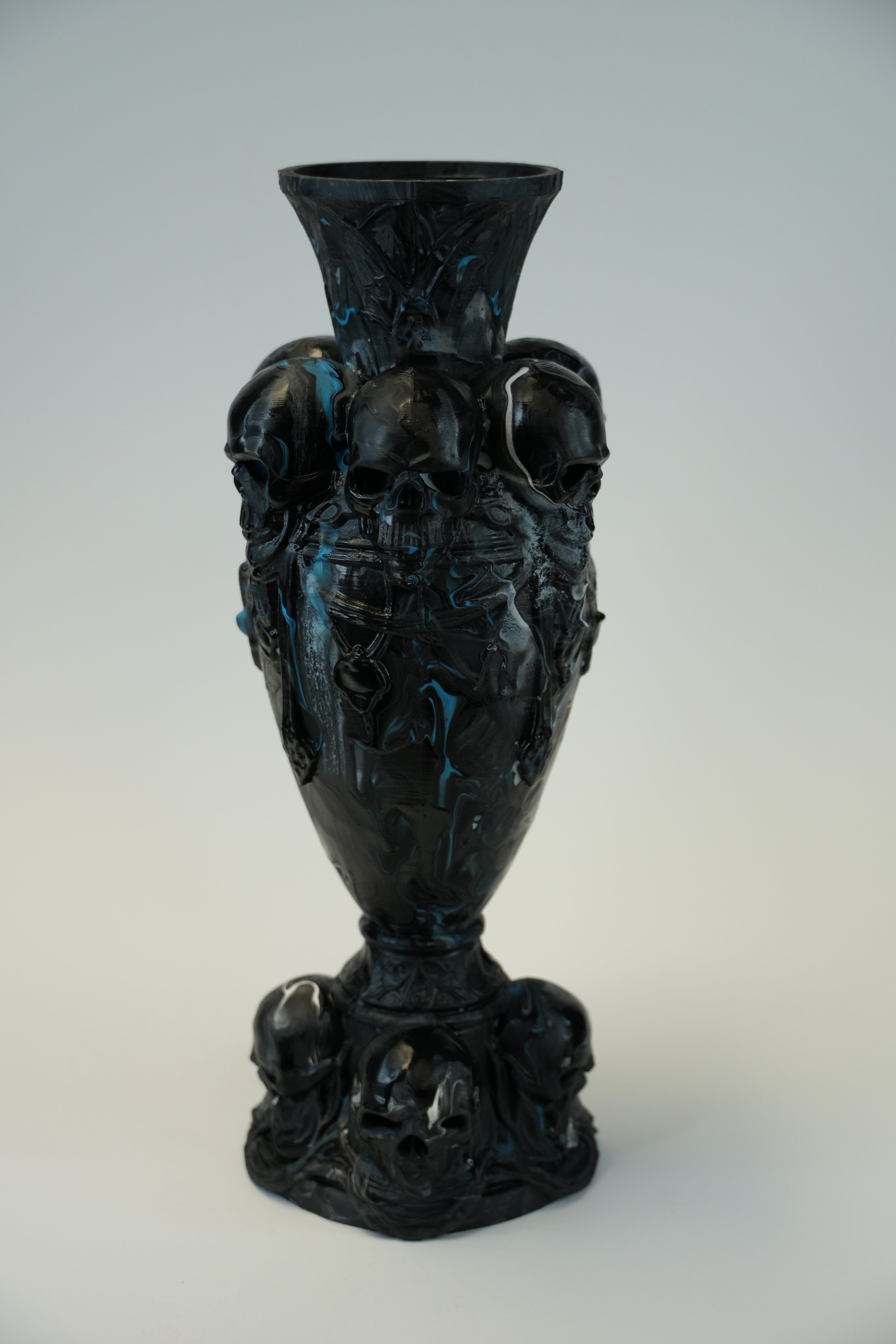 A black gothic-style ceramic vase decorated with sculpted skulls and dark blue marbled accents, set against a neutral light gray background.