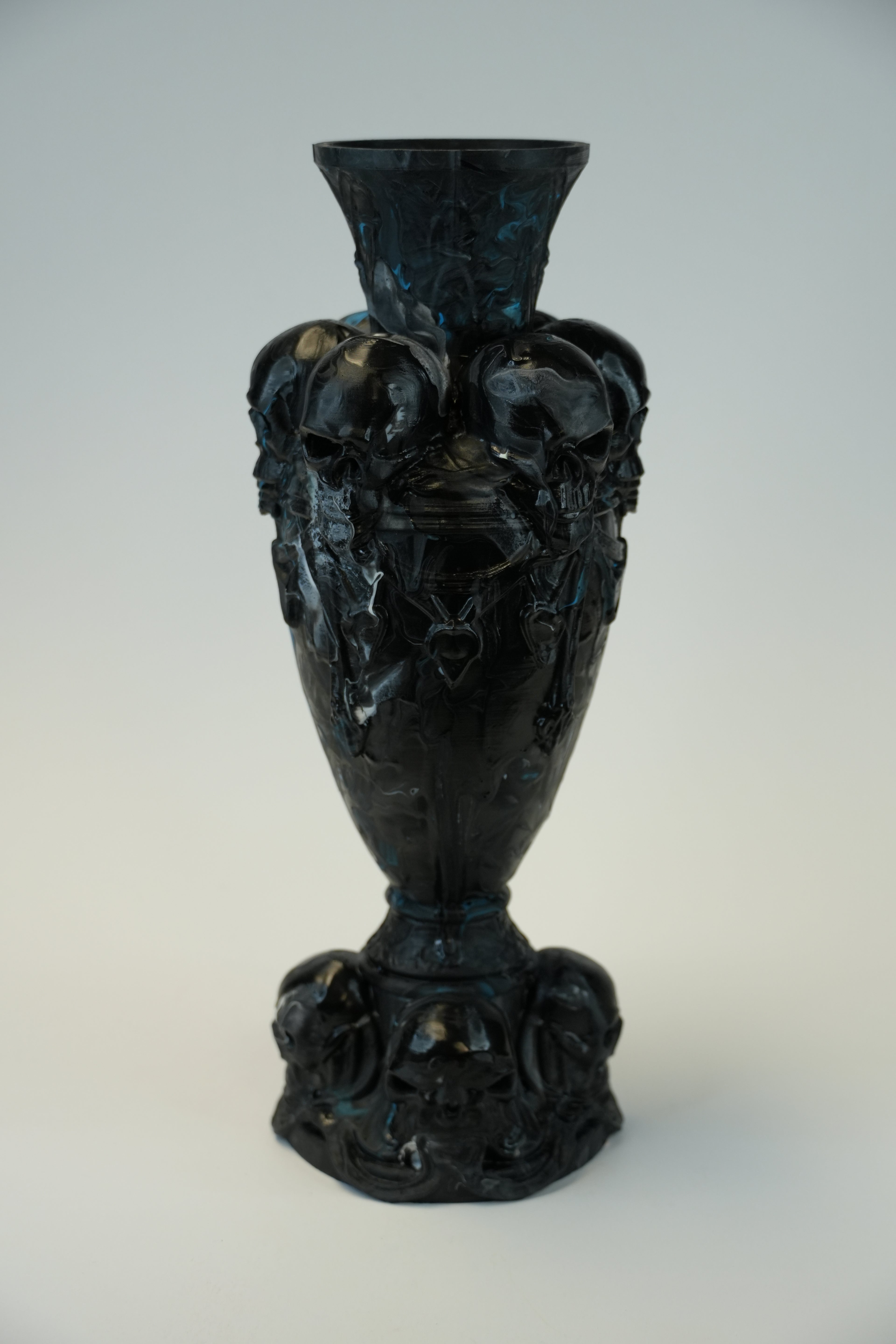 A dark, ornate gothic-style vase featuring sculpted skull details and a flared rim, photographed against a neutral light gray background.