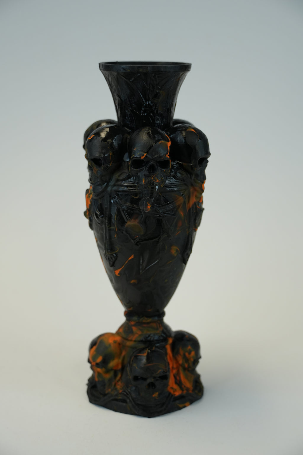 A dark, ornate sculptural vase with skull motifs and marbled orange-brown accents, photographed against a plain light gray studio background.