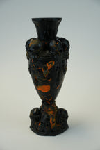 A black ceramic vase with a flared rim, sculpted side details, and a pedestal base, decorated with swirling orange and amber marbled patterns, set against a light gray background.