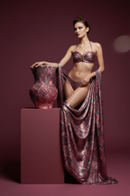 Woman in a patterned bikini and cover-up standing next to a decorative vase on a pink platform.