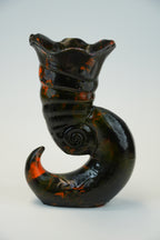 A sculptural ceramic vase with a glossy black glaze and vibrant orange and brown marbled accents, photographed against a neutral light gray background.