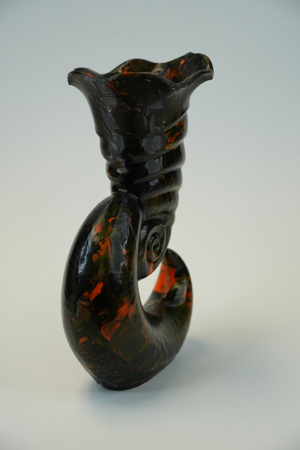 A sculptural ceramic vase with a glossy black and terracotta orange marbled pattern, featuring a flared rim and a curved body, photographed on a neutral light gray background.