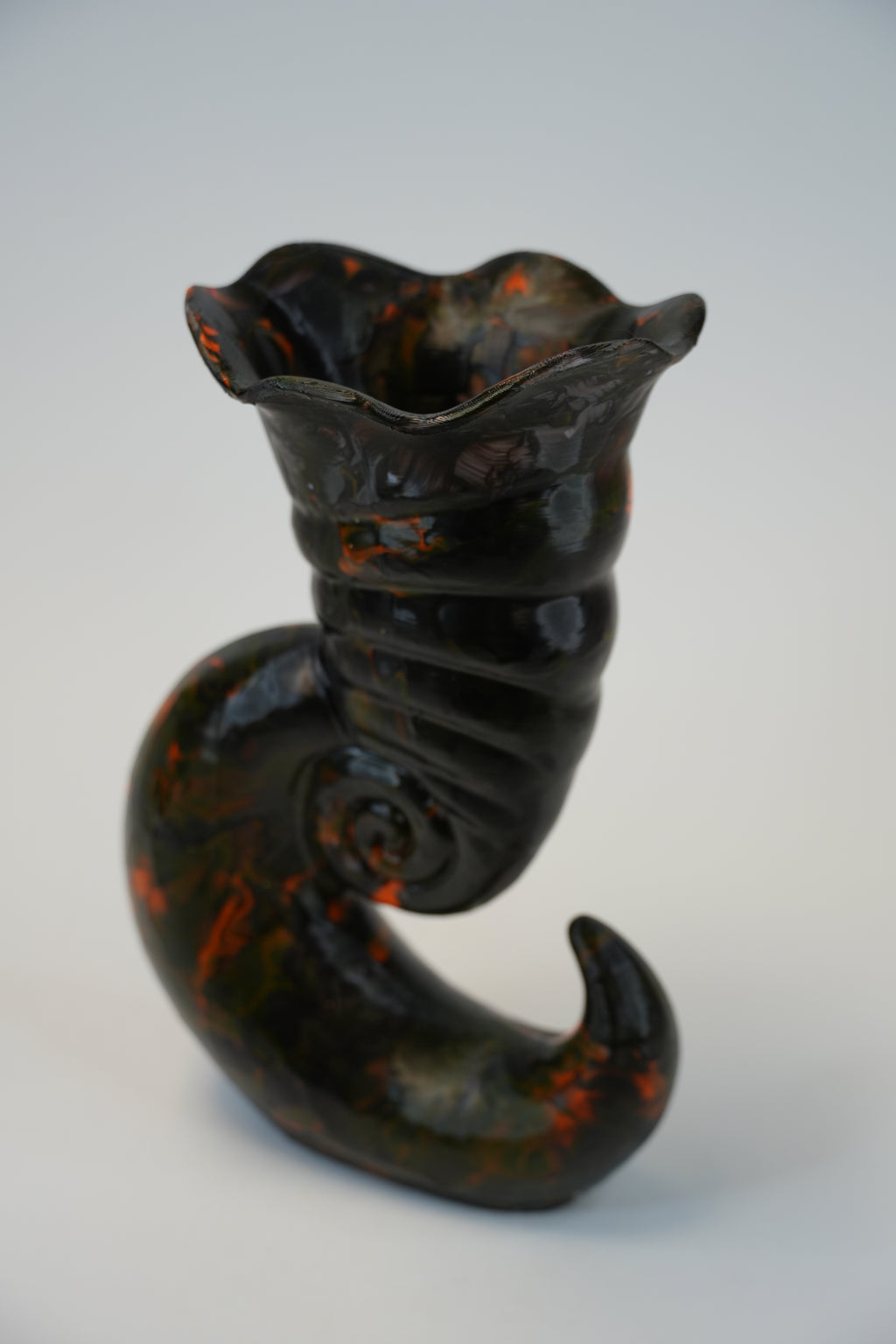 A sculptural ceramic vase with a dark glossy finish and orange marbled accents, shaped like a curled spiral shell or horn, photographed on a light gray background.