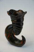 A sculptural ceramic vase with a dark glossy finish and orange marbled accents, shaped like a curled spiral shell or horn, photographed on a light gray background.