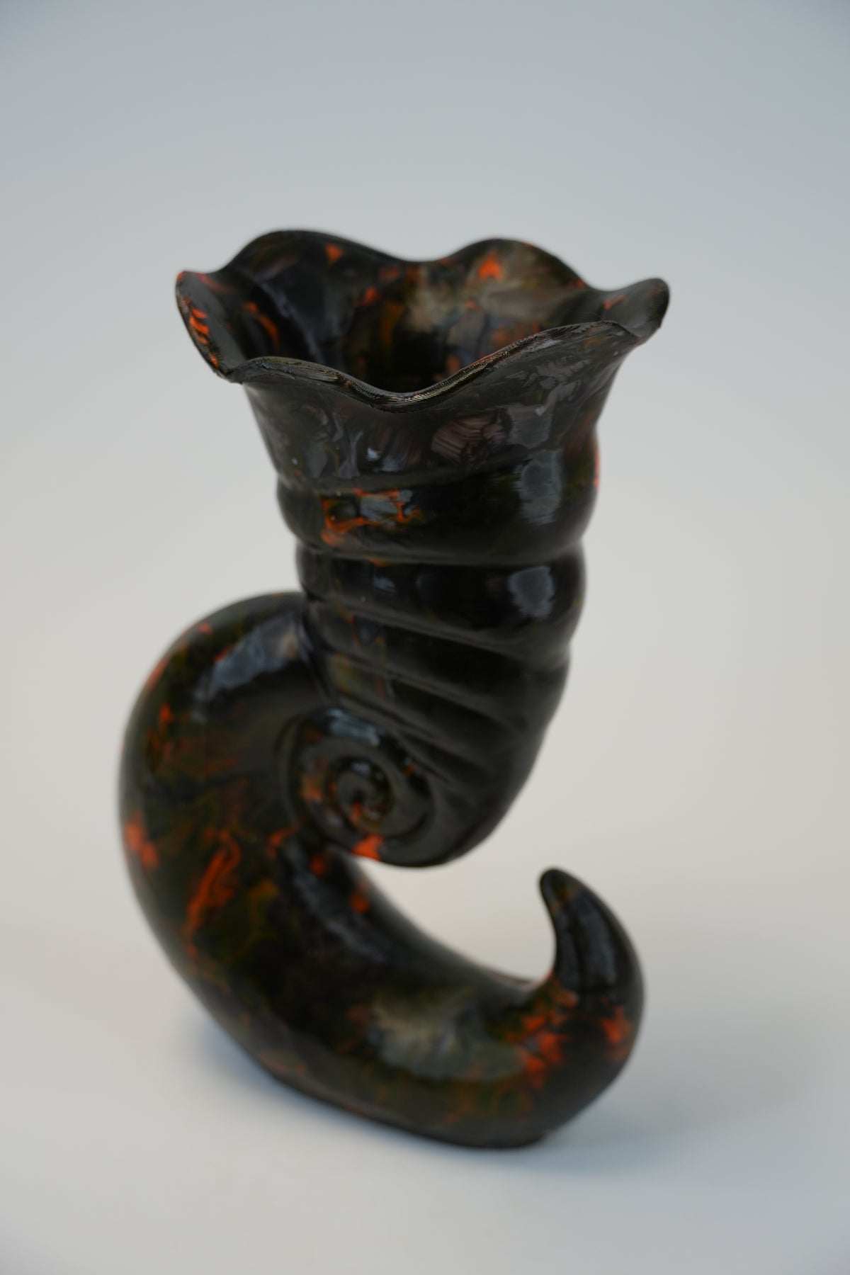 A sculptural ceramic vase with a dark glossy finish and orange marbled accents, shaped like a curled spiral shell or horn, photographed on a light gray background.