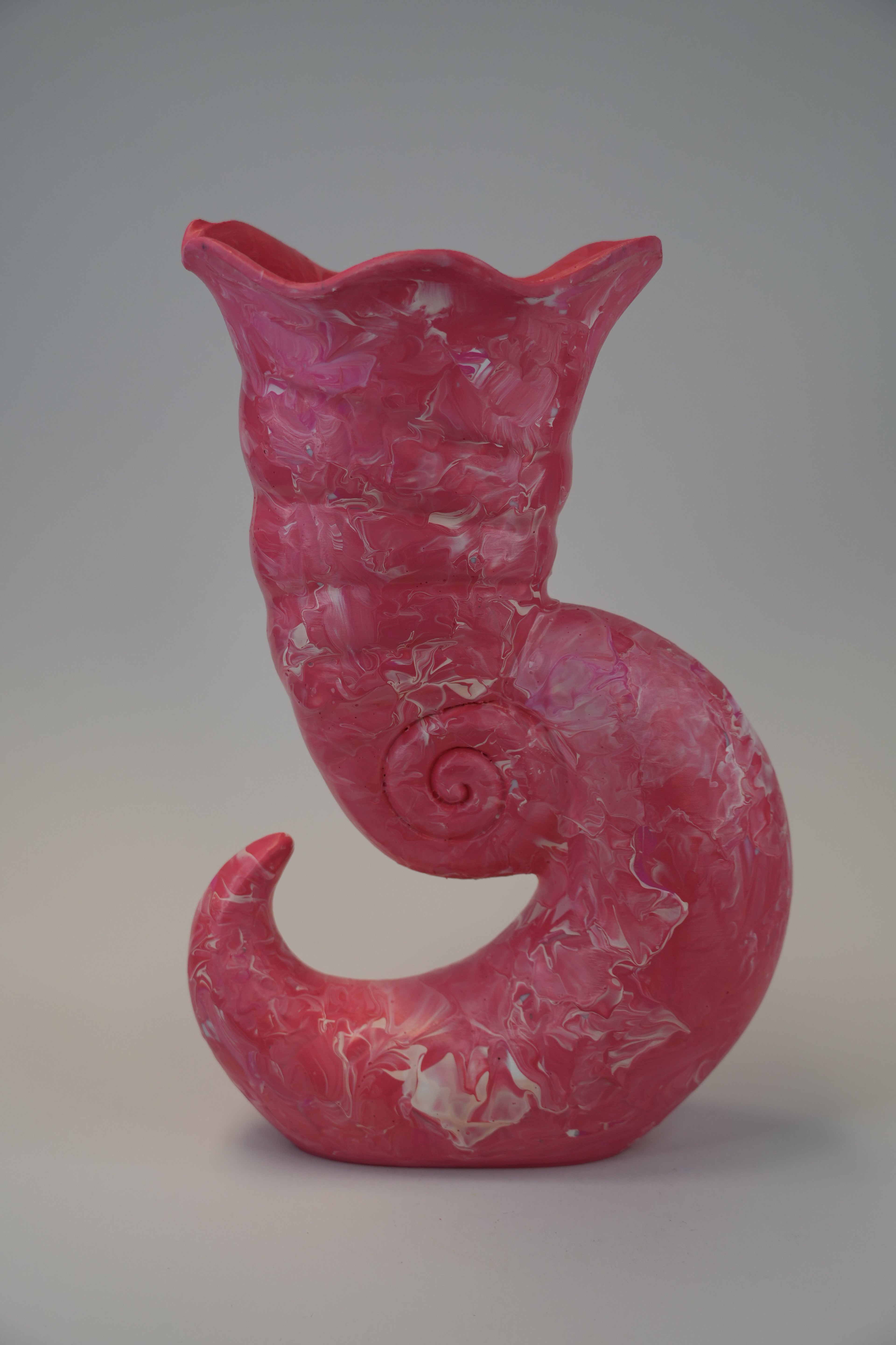A sculptural ceramic vase shaped like a curled shell or spiral shell, in a glossy pink glaze with white marbled veining, photographed against a neutral gray studio background.