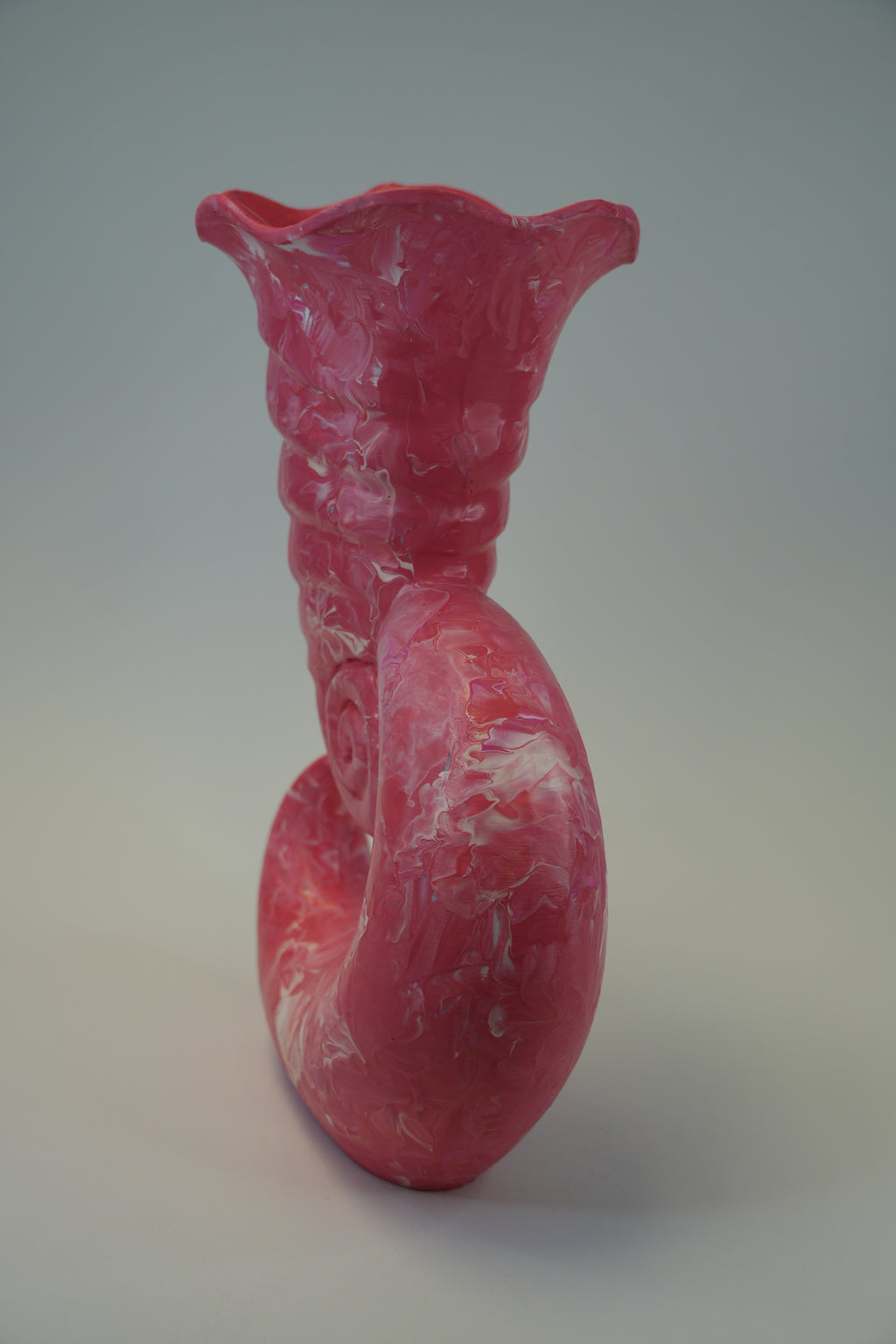 A sculptural ceramic vase with a curved, coiled form and a flared rim, featuring a pink and white marbled pattern, photographed against a neutral gray background.