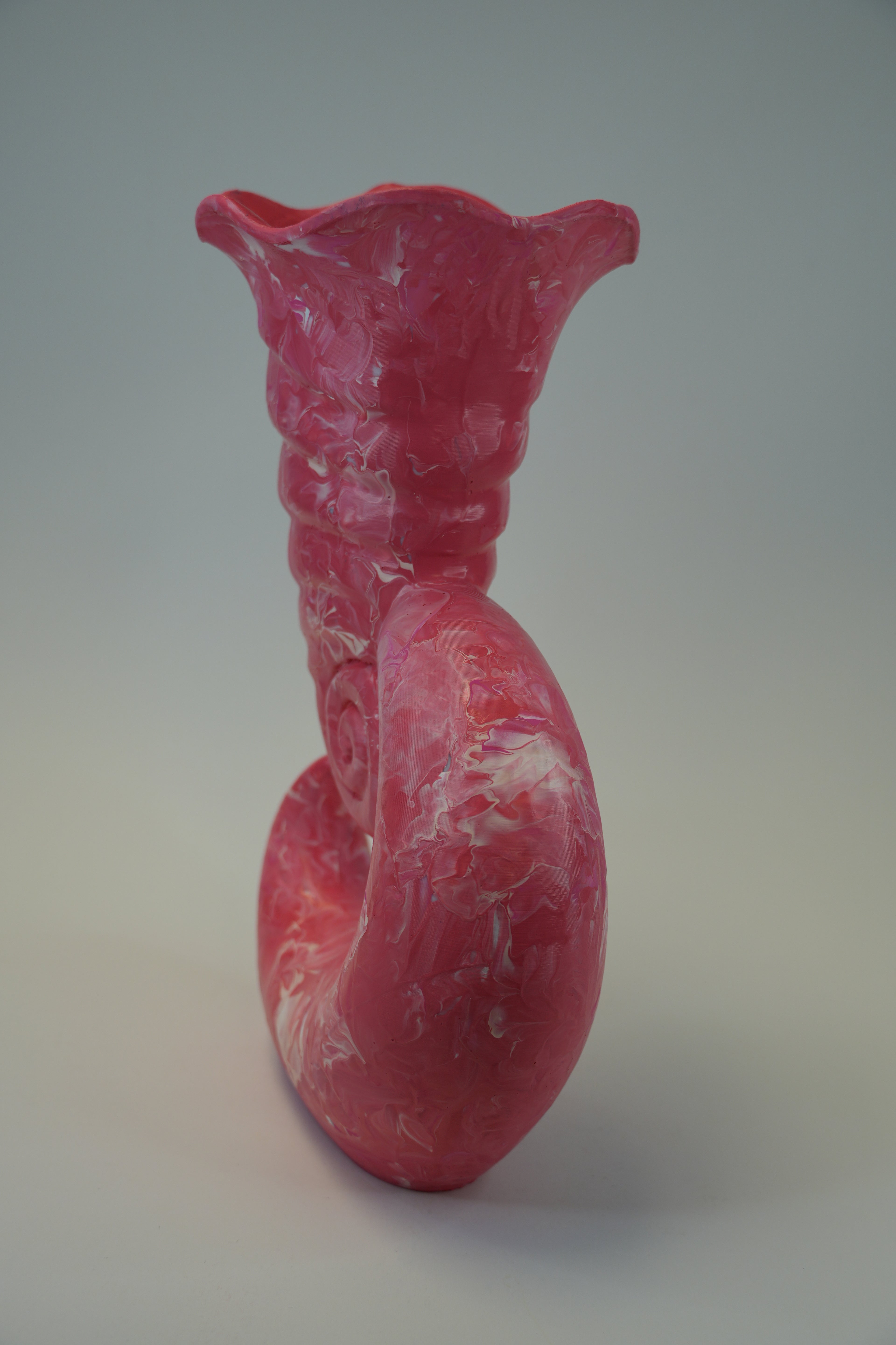 A sculptural ceramic vase with a curved, coiled form and a flared rim, featuring a pink and white marbled pattern, photographed against a neutral gray background.