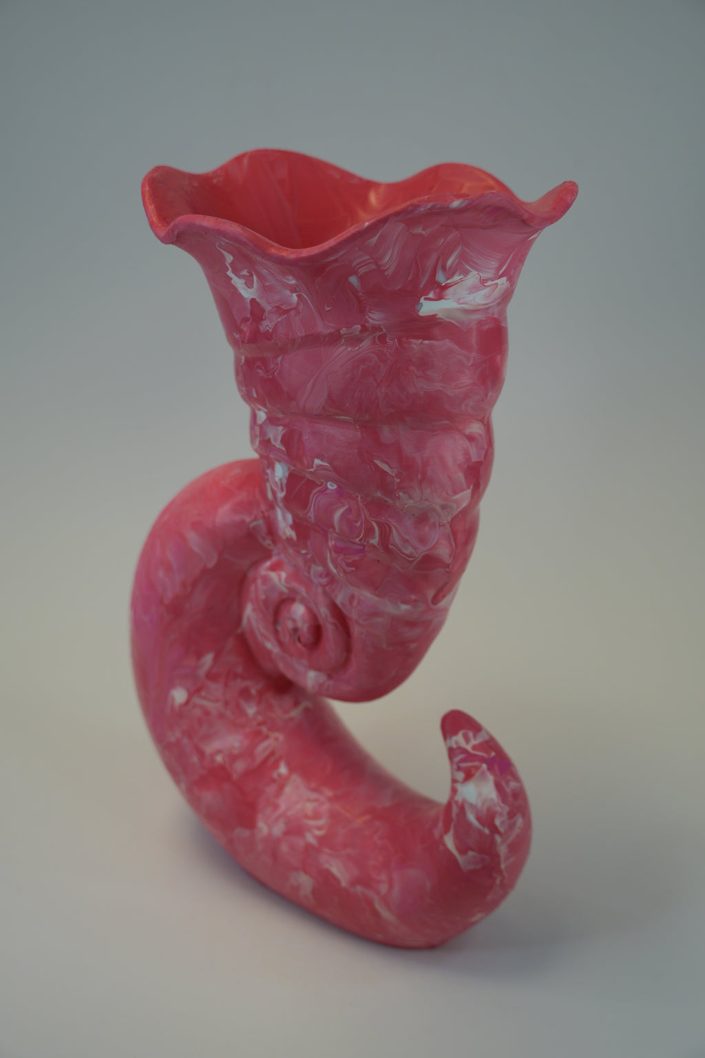A sculptural pink ceramic vase with a flared top and a curved, spiral-like base, shown on a neutral light gray studio background.