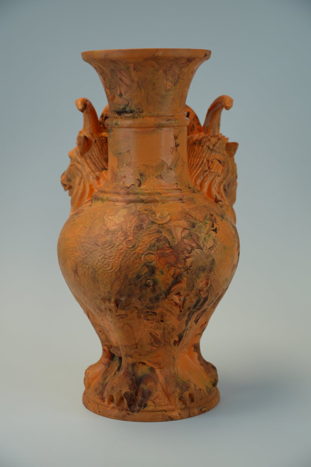An orange glazed ceramic vase with carved lion heads on the sides and a wide flared rim, shown against a light gray studio background.